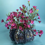 A decorative dark metallic vase with a textured, hammered finish, filled with a dense arrangement of small pink flowers with green stems, set against a solid light blue background.