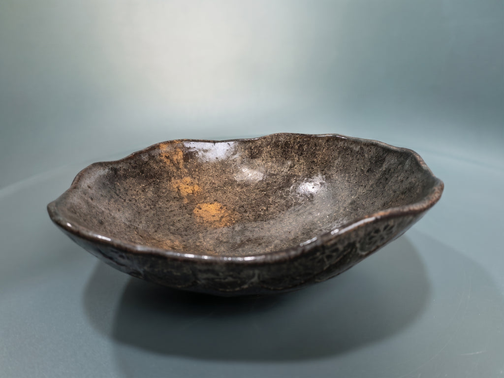 A handmade ceramic bowl with a dark brown and black textured glaze, featuring a natural organic scalloped rim and subtle orange-brown inclusions inside, set against a smooth gray background.
