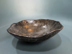 A handmade ceramic bowl with a dark brown and black textured glaze, featuring a natural organic scalloped rim and subtle orange-brown inclusions inside, set against a smooth gray background.