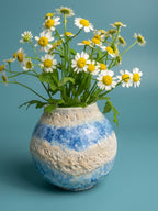 A ceramic vase with blue and beige marbled patterns, filled with white daisies that have yellow centers and green stems, set against a solid teal-blue background.