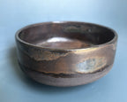 A handmade ceramic bowl with a rustic, hand-painted finish, featuring a dark brown base with metallic bronze and iridescent blue-green glazes, set against a soft light blue background.