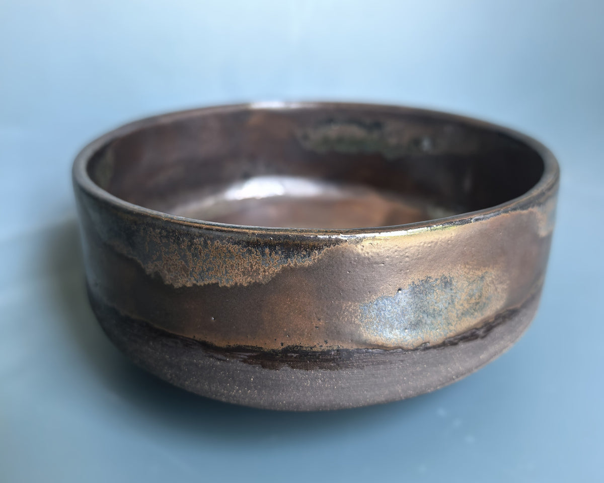 A handmade ceramic bowl with a rustic, hand-painted finish, featuring a dark brown base with metallic bronze and iridescent blue-green glazes, set against a soft light blue background.
