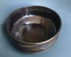 A handmade ceramic bowl with a rustic, hand-painted finish in dark brown, bronze, and green glazes, shown on a light blue-gray background.