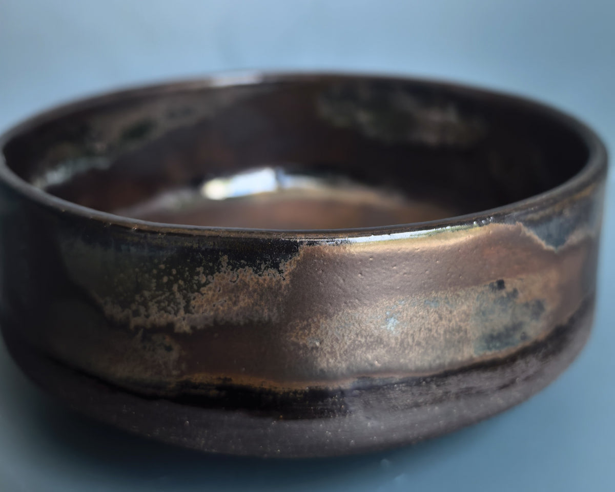 A handmade ceramic bowl with a rustic, hand-painted finish, featuring a dark brown base with metallic bronze and black textured glazes, set against a solid blue-gray background.