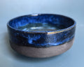 A handmade ceramic bowl with a glossy deep blue glazed upper half and a matte, speckled dark gray-brown lower half, set against a soft light blue background.