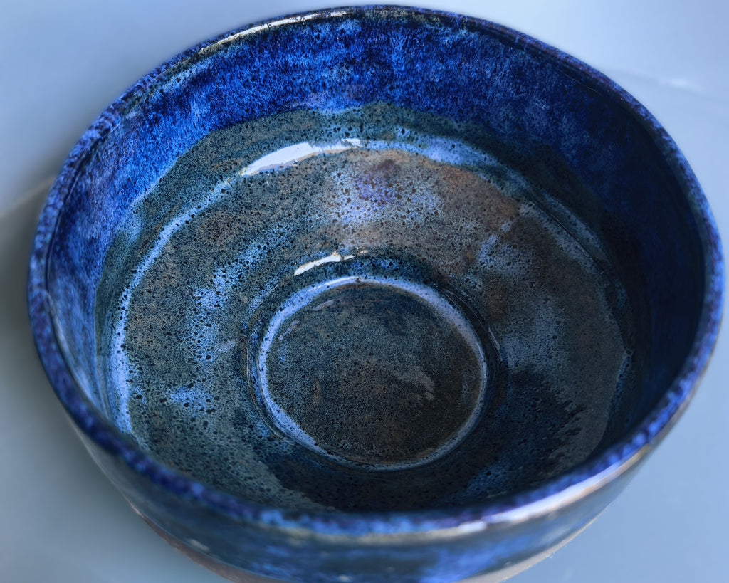 A handmade ceramic bowl with a glossy, textured glaze featuring a deep blue outer rim and a mottled, speckled interior in shades of blue, gray, and brown.