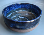 A handmade ceramic bowl with a glossy deep blue glaze on the inside and rim, transitioning to a matte, speckled brown clay base on the outside, shown on a light blue-gray background.