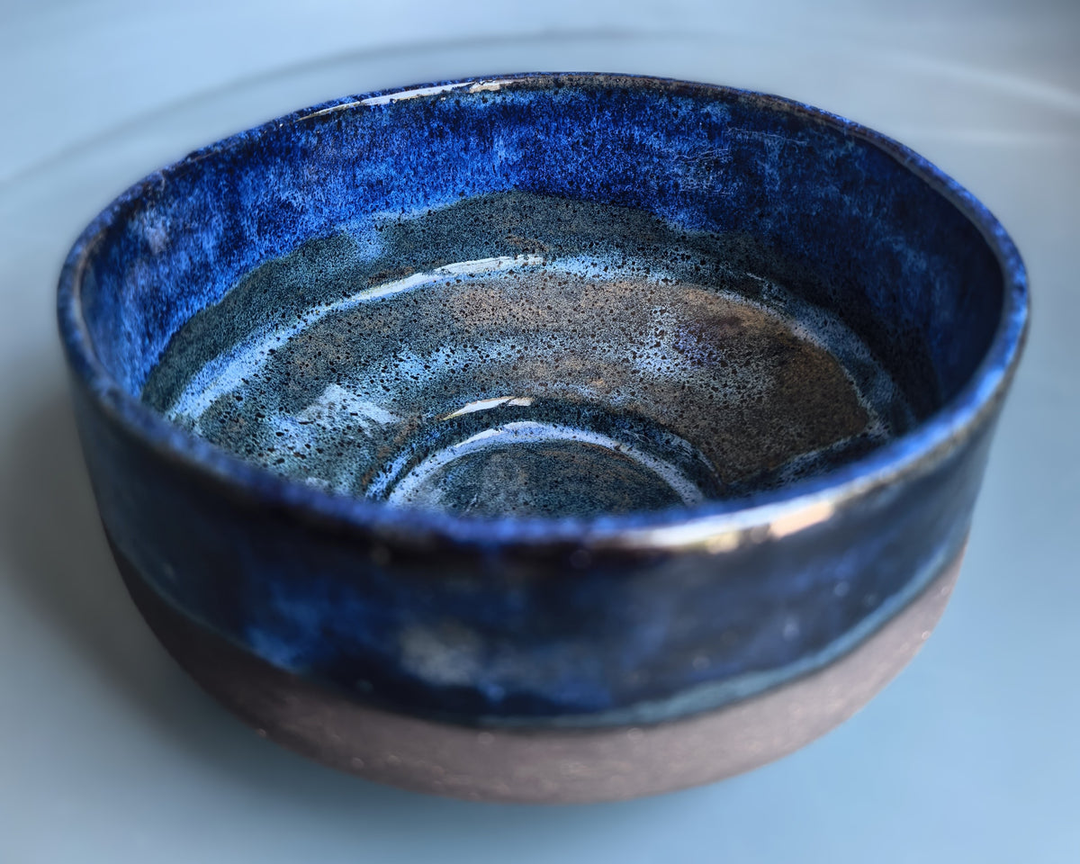 A handmade ceramic bowl with a glossy deep blue glaze on the inside and rim, transitioning to a matte, speckled brown clay base on the outside, shown on a light blue-gray background.