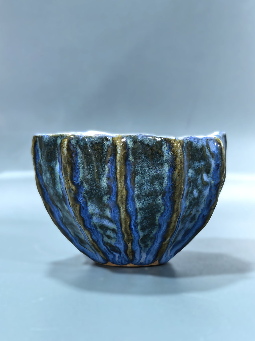 A handmade ceramic bowl with a textured, ribbed exterior featuring a glossy glaze of deep blue, dark green, and metallic gold stripes, set against a neutral gray background.