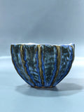 A handmade ceramic bowl with a textured, ribbed exterior featuring a glossy glaze of deep blue, dark green, and metallic gold stripes, set against a neutral gray background.
