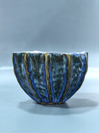 A handmade ceramic bowl with a textured, ribbed exterior featuring a glossy glaze of deep blue, dark green, and metallic gold stripes, set against a neutral gray background.