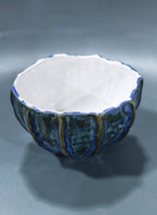 A handmade ceramic bowl with a textured, ribbed exterior in blue and brown glazes, featuring a smooth white interior, set against a dark gray background.