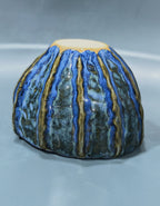 A handmade ceramic bowl with a textured, ribbed surface featuring vertical blue, green, and gold glazes, photographed on a neutral gray background.