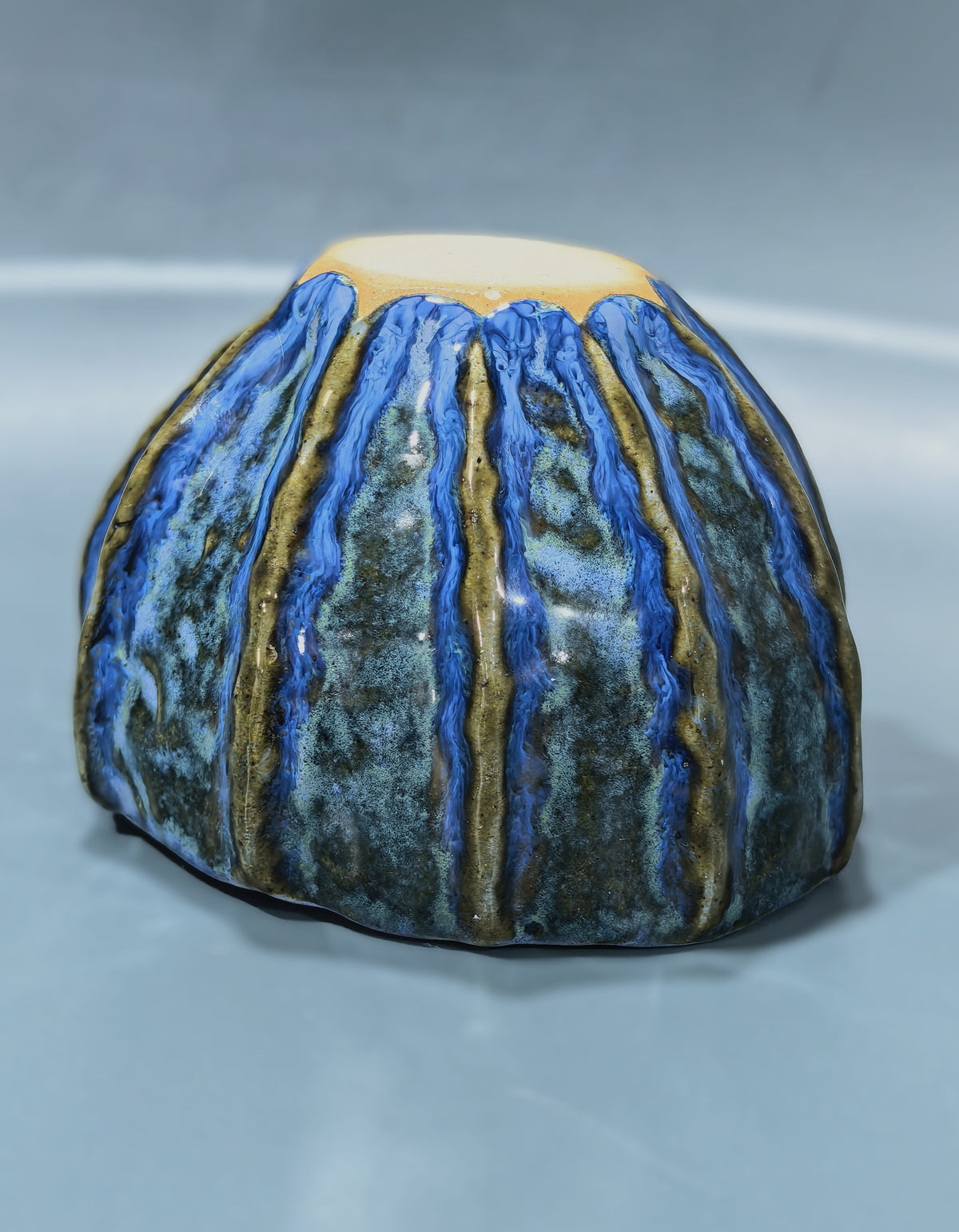A handmade ceramic bowl with a textured, ribbed surface featuring vertical blue, green, and gold glazes, photographed on a neutral gray background.