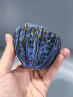 A handmade ceramic bowl with a textured, ribbed design, held in a person's hand. The bowl features a glossy blue and black glaze with metallic gold lines separating the vertical ridges, set against a neutral gray background.