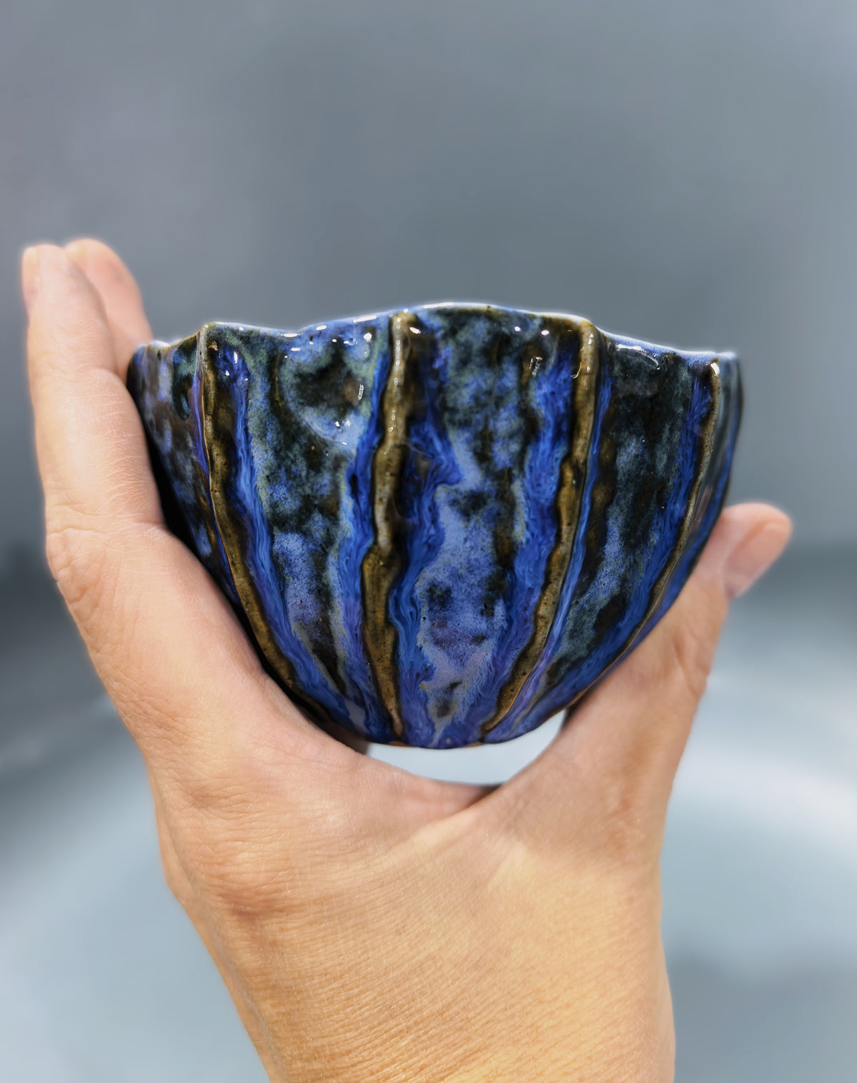 A hand holding a small handmade ceramic bowl with a blue, black, and gold marbled glaze against a soft gray background.