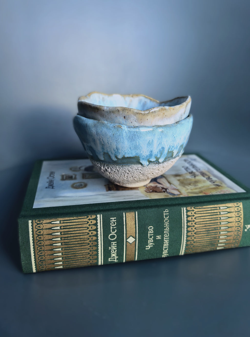 Two stacked handmade ceramic bowls with a blue and beige glaze, displayed on top of a vintage green book against a dark gray background.