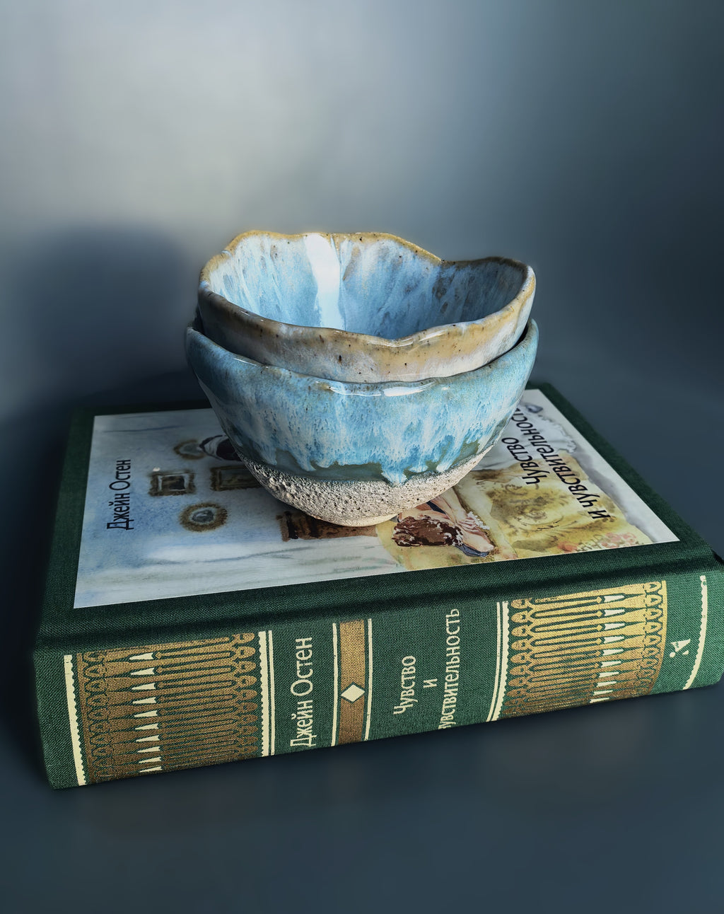 Two stacked handmade ceramic bowls with a blue and beige glaze, displayed on top of a vintage green book against a dark gray background.