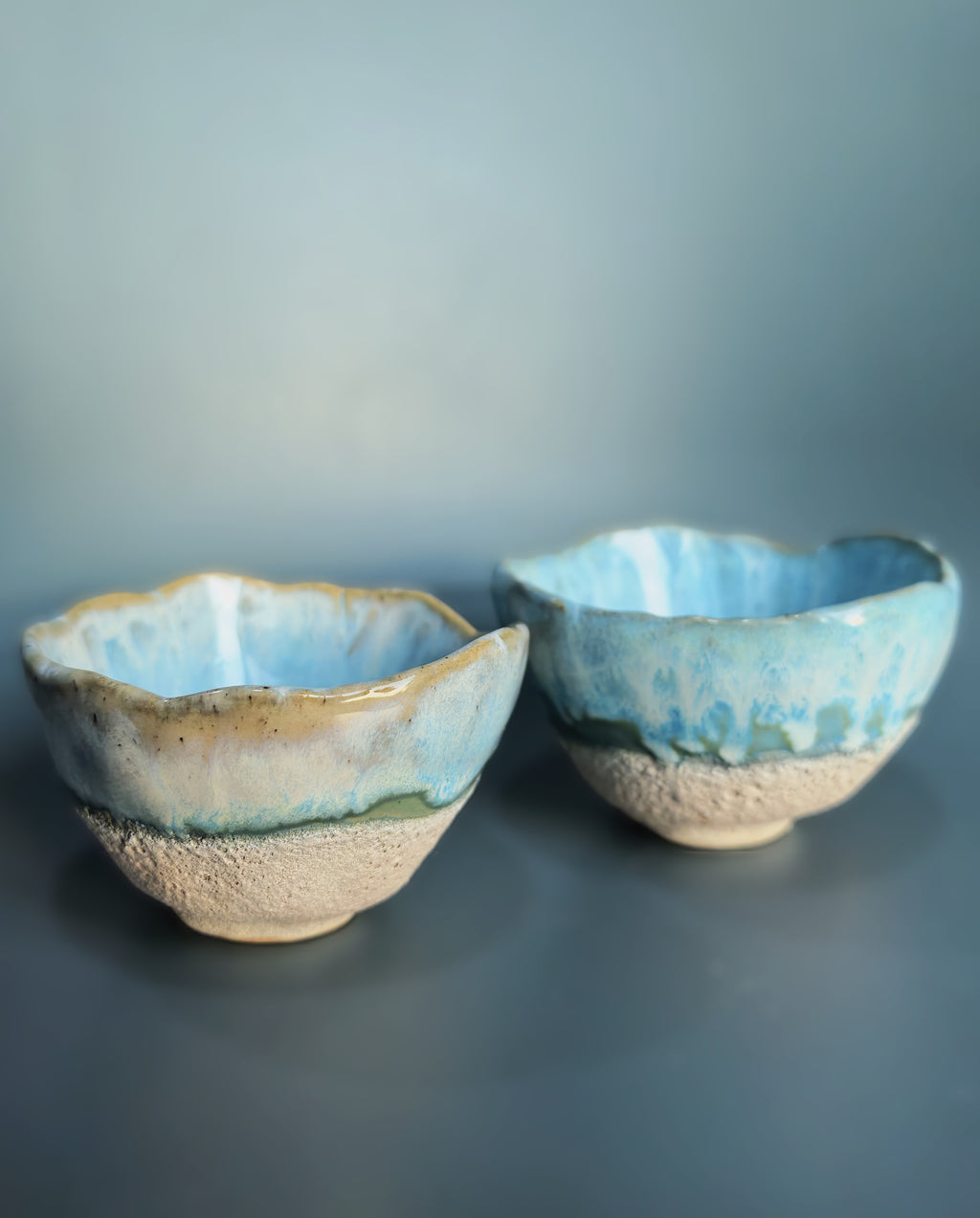 Two handmade ceramic bowls with a light blue and white glaze and a textured beige base, set against a dark blue-gray background.