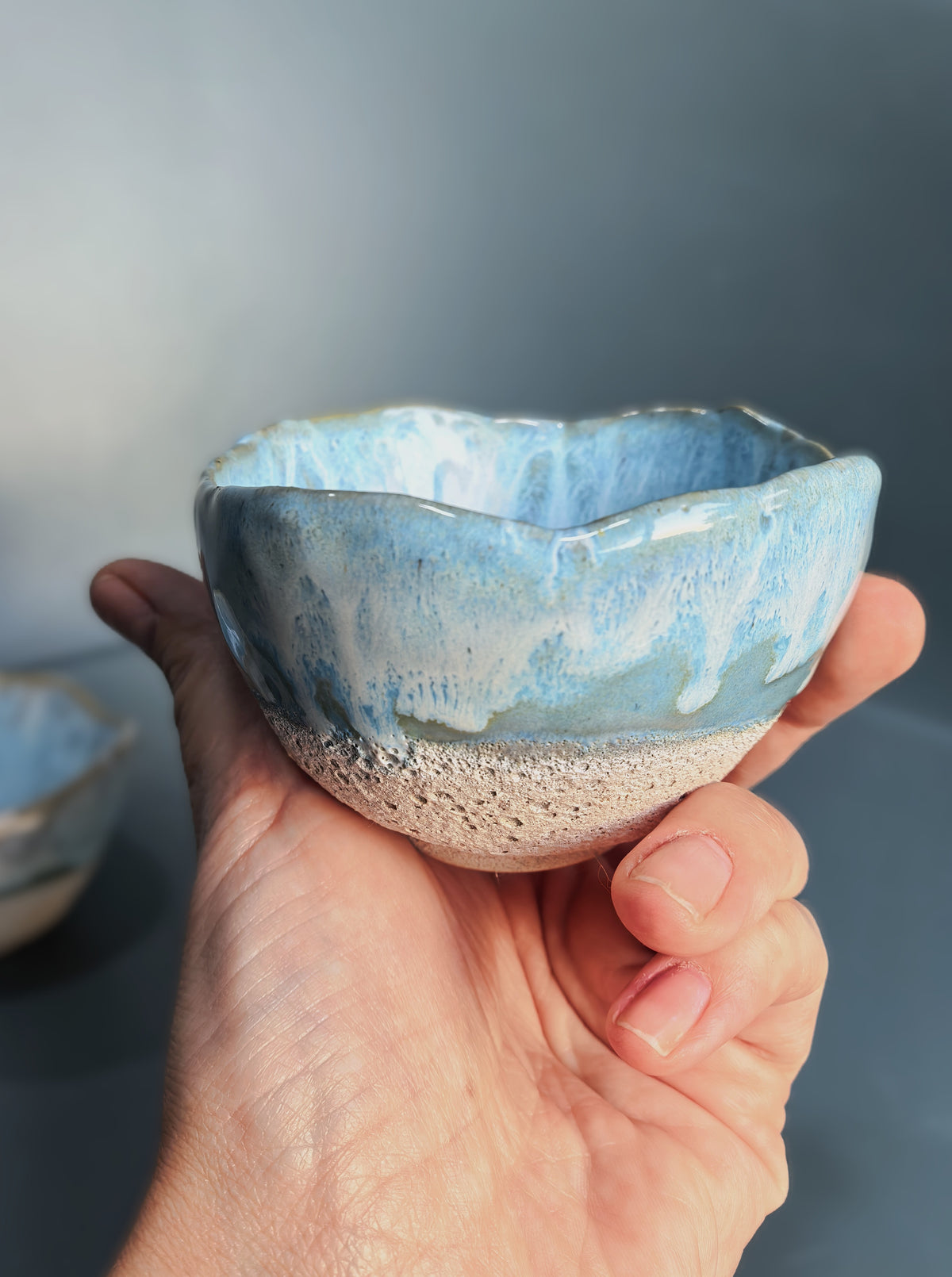 A hand holding a small handmade ceramic bowl with a blue and white marbled glaze on the upper half and a rough, speckled beige base on the lower half.