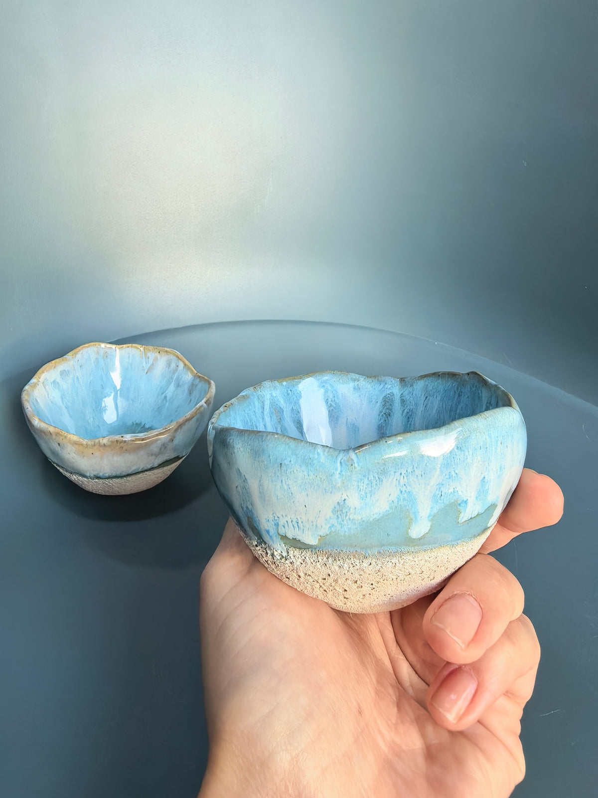 A hand holds a small handmade ceramic bowl with a blue and white glaze and a speckled beige base, with a second similar bowl blurred in the background against a blue-gray backdrop.