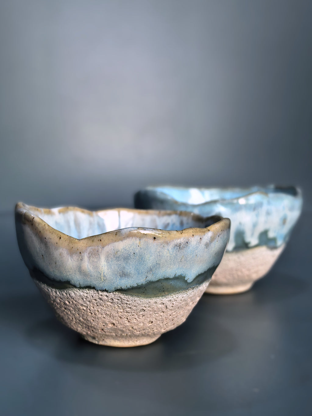 Two handmade ceramic tea bowls with a textured beige base and a glossy blue-green glaze, shown on a dark gray background.