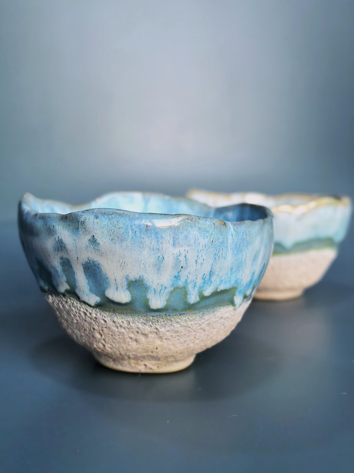 Two handmade ceramic tea bowls with a blue and white dripping glaze and a textured beige base, shown on a dark gray surface against a blue-gray background.