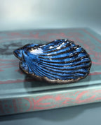 A small shell-shaped dish with a glossy blue and black striped glaze, resting on a vintage book with a blue-gray background.