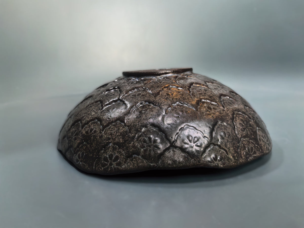 A dark brown ceramic bowl with a textured, embossed floral pattern and a small top handle, photographed against a neutral gray background.