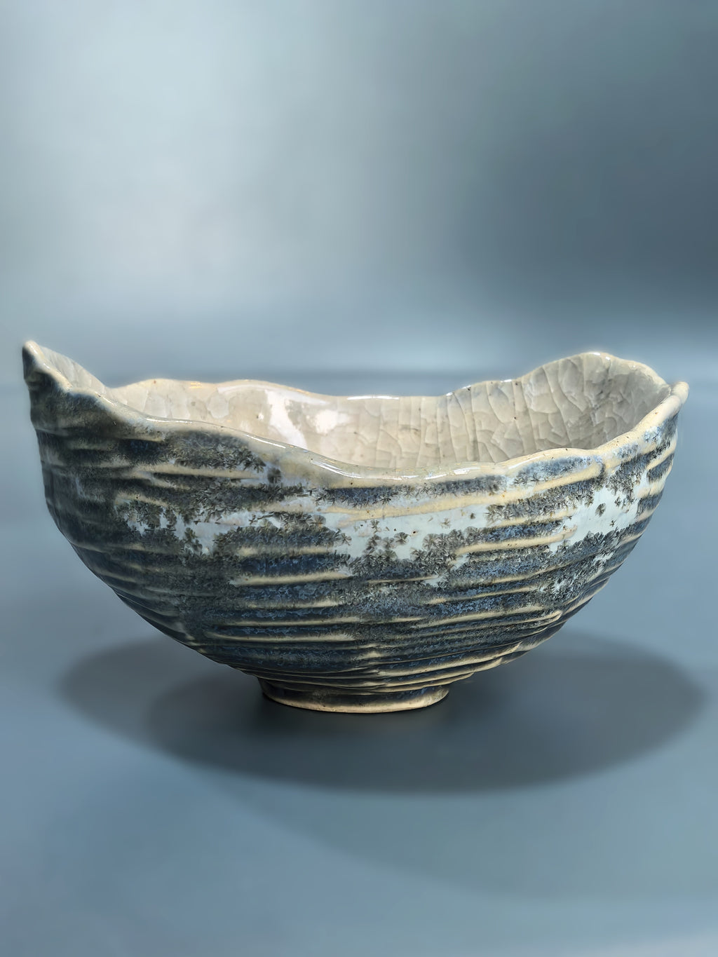 A handmade ceramic bowl with a textured, ribbed exterior and a cracked, glossy interior, shown on a neutral gray background.
