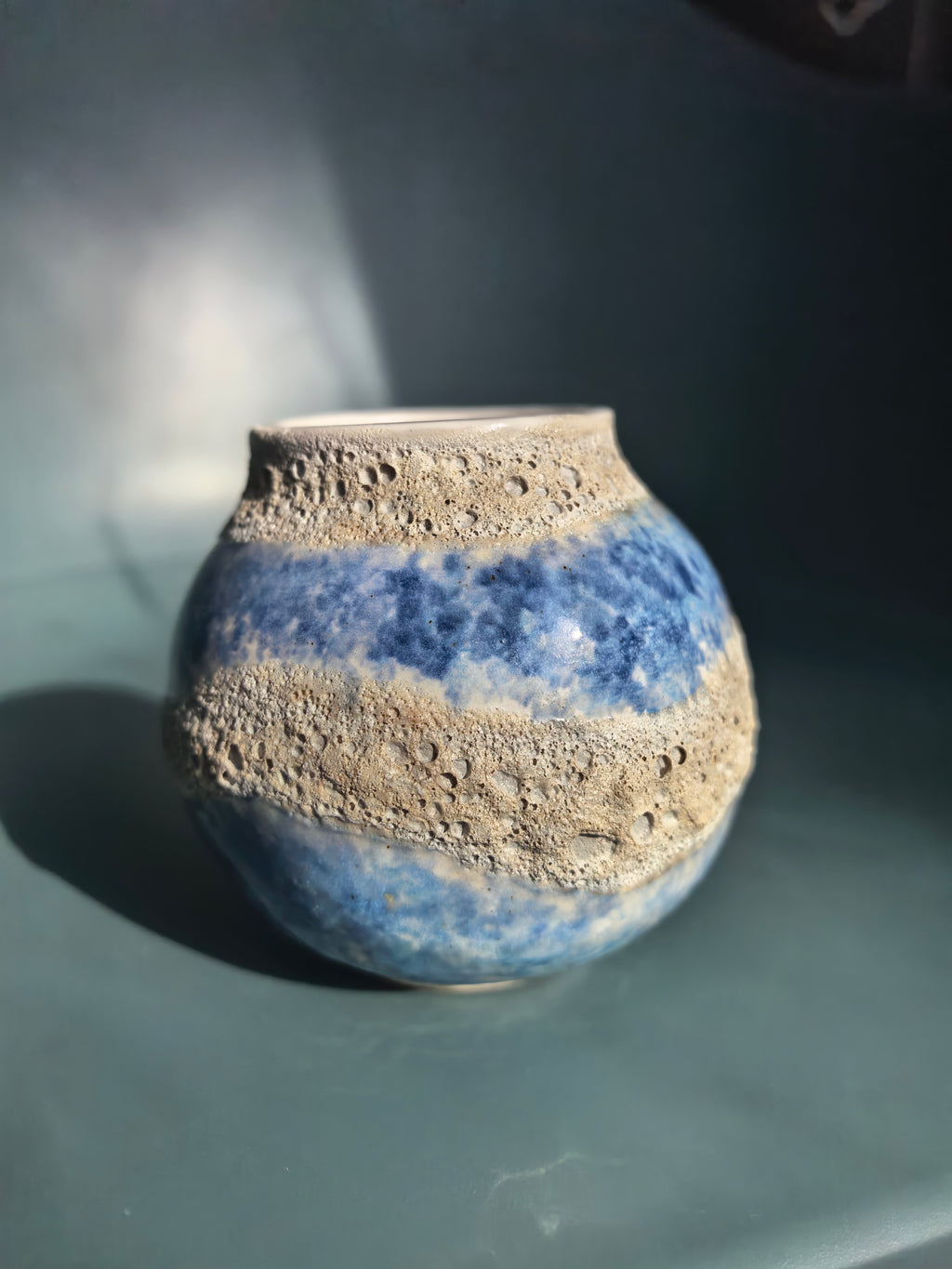 A small round ceramic vase with a blue and white marbled pattern and rough, textured beige bands, photographed on a dark teal surface with dramatic side lighting.