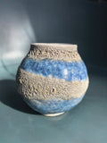 A handmade ceramic vase with a textured, porous sandy beige finish and horizontal bands of glossy cobalt blue glaze, set against a dark blue-gray background.