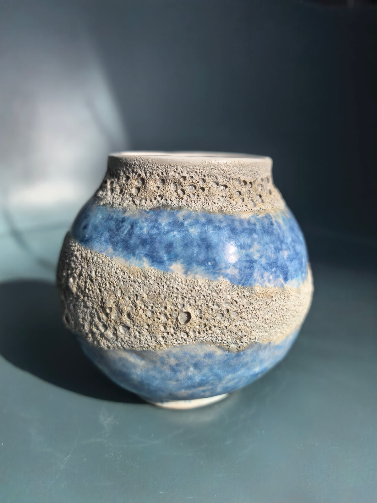 A handmade ceramic vase with a textured, porous sandy beige finish and horizontal bands of glossy cobalt blue glaze, set against a dark blue-gray background.