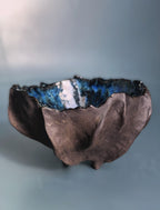 An organic-shaped ceramic bowl with a dark, textured exterior and a glossy blue, white, and black speckled interior, set against a soft blue-gray background.