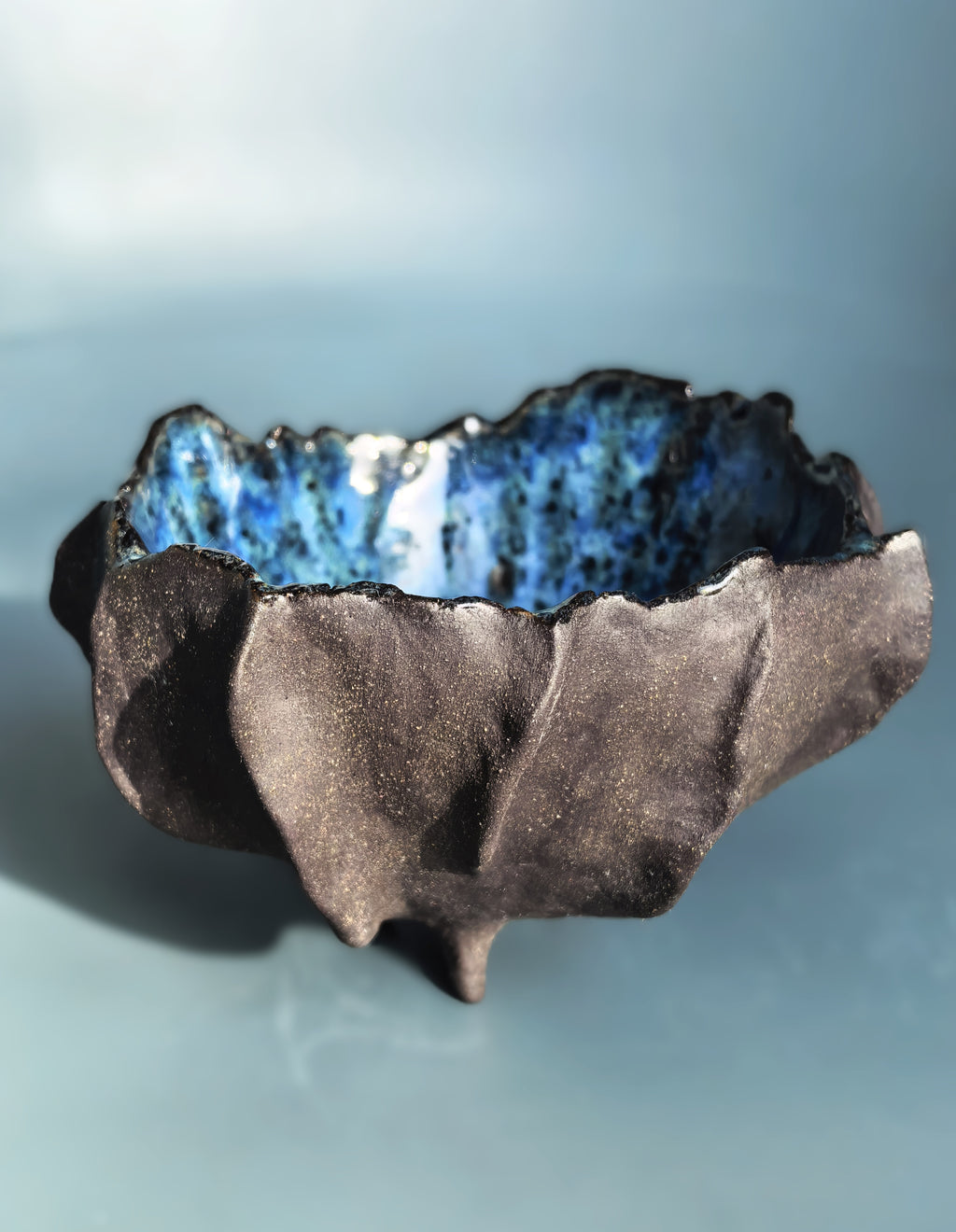 A handmade ceramic bowl with a dark, textured, organic-looking exterior and a vibrant blue glazed interior, set against a soft blue background.