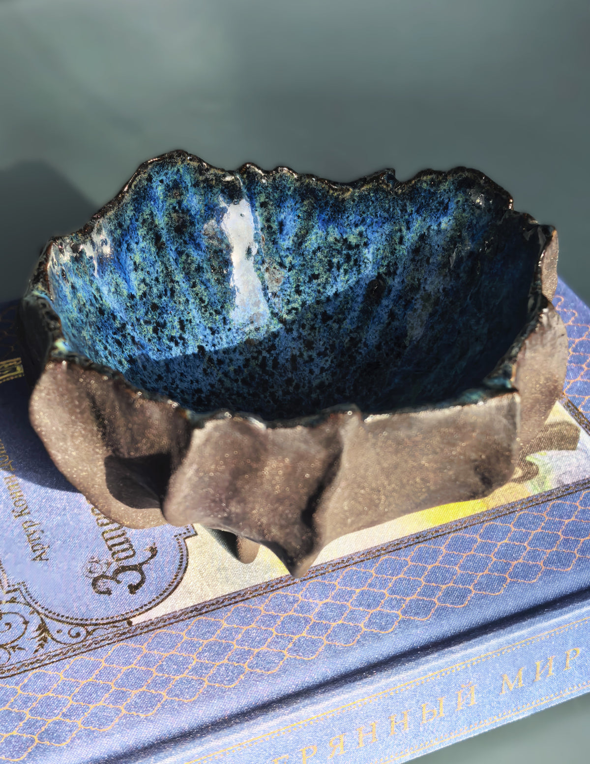 A handmade ceramic bowl with a textured, organic shape, featuring a glossy dark blue and black glazed interior and a rustic brown exterior, resting on top of a vintage blue book.