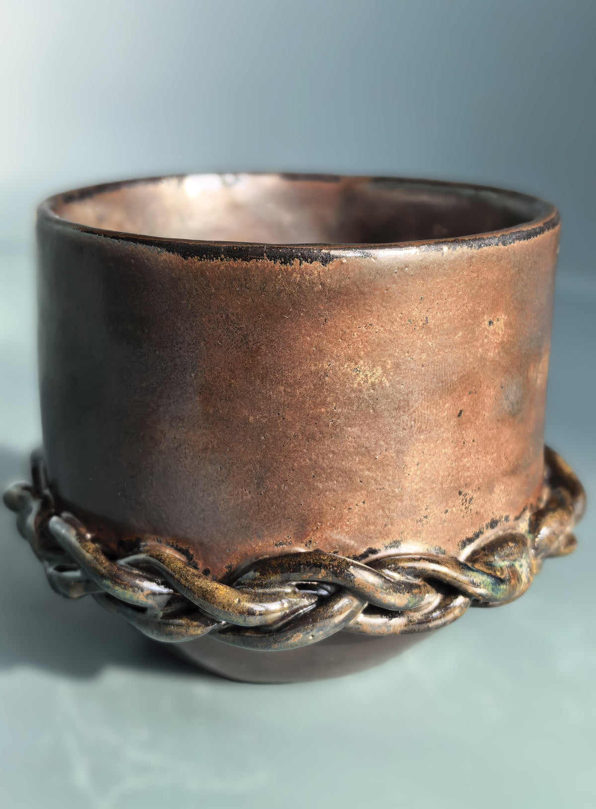 A handmade ceramic cup with a rustic copper-brown glaze and a braided metallic band around the base, shown on a soft blue-gray background.