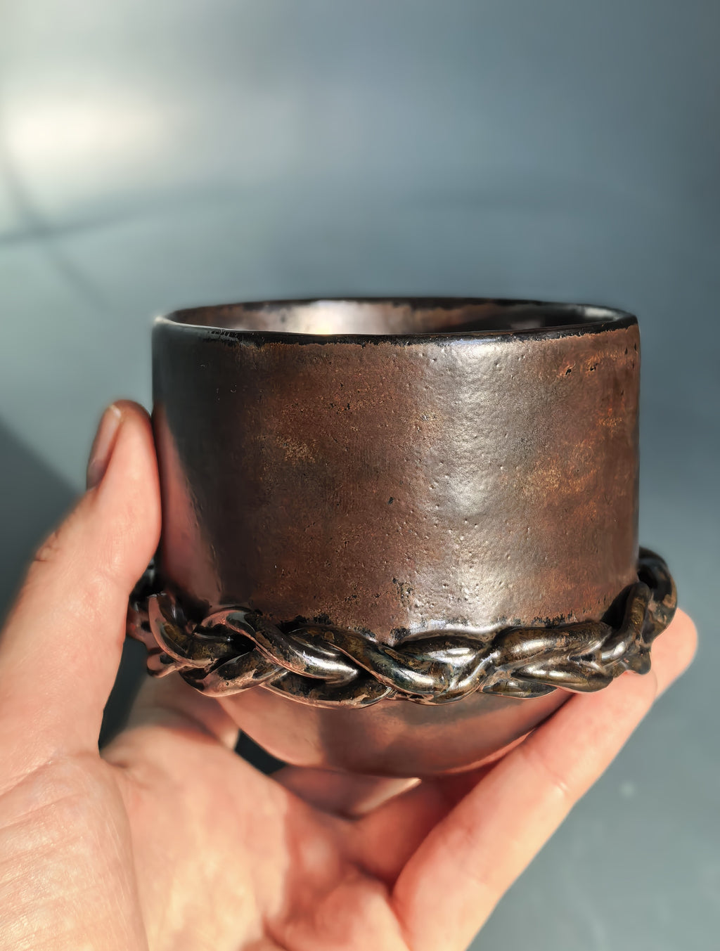 A hand holding a rustic dark brown ceramic cup with a braided metallic band around the lower half, set against a soft blue-gray background.