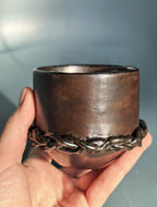A hand holding a rustic dark brown ceramic cup with a braided metallic band around the lower half, set against a soft blue-gray background.