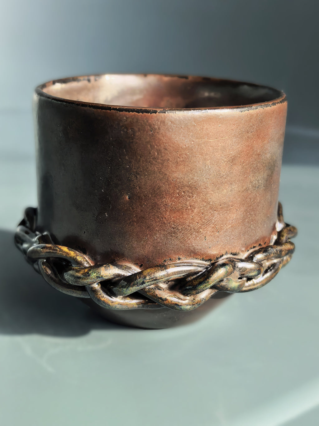 A rustic handmade copper-colored ceramic cup with a thick braided metal chain wrapped around its base, set against a neutral gray background.