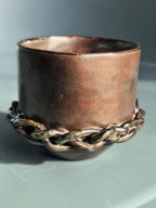 A rustic handmade copper-colored ceramic cup with a thick braided metal chain wrapped around its base, set against a neutral gray background.