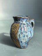 A handmade ceramic pitcher with a rounded body, a small side handle, and a textured glaze featuring light blue, dark blue, and metallic gold-brown sections, set against a neutral gray background.