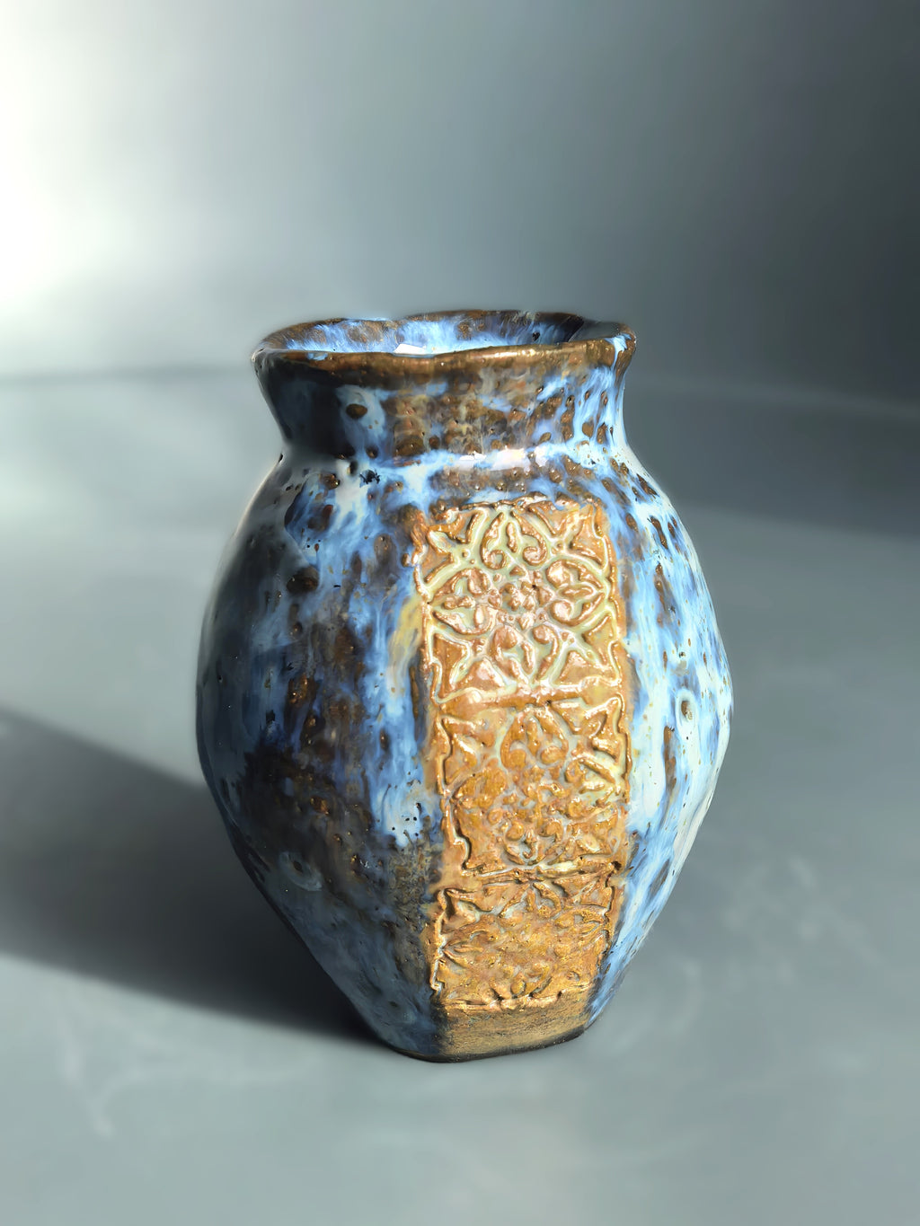 A handmade ceramic vase with a rounded body, flared rim, and a textured gold-toned panel decorated with an intricate floral pattern, covered in a mottled blue and brown glaze.