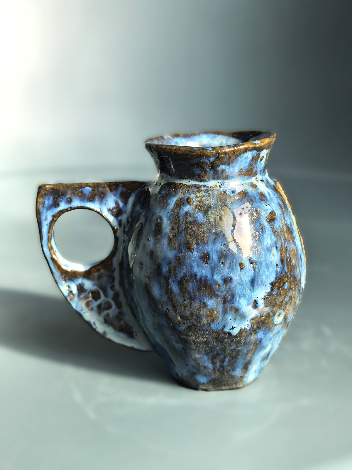 A handmade ceramic pitcher with a rounded body, a loop handle, and a short spout, decorated with a mottled blue and brown glaze on a neutral gray background.