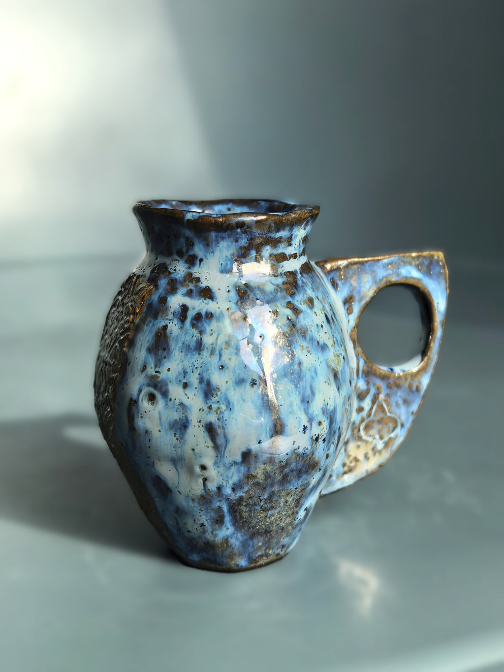 A handmade ceramic pitcher with a rounded body, a small spout, and a loop handle, decorated with a mottled blue, white, and brown glaze on a gray background.