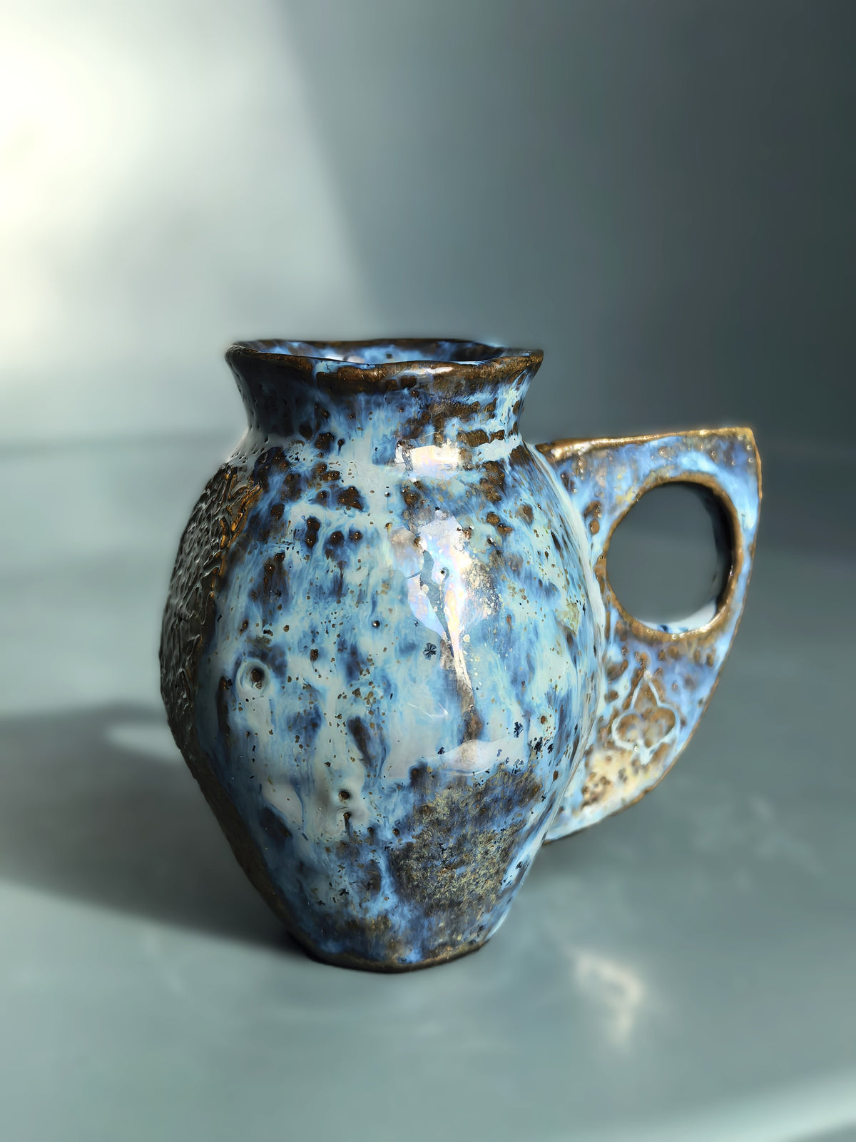 A handmade ceramic pitcher with a rounded body, a small spout, and a loop handle, decorated with a mottled blue, white, and brown glaze on a gray background.