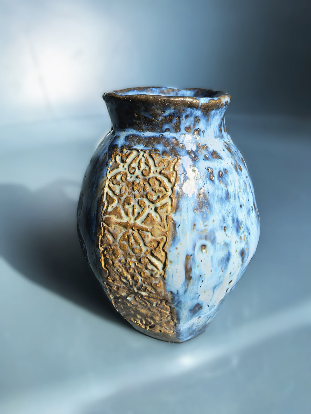 A small handmade ceramic vase with a rounded bulbous body, featuring a split glaze design of textured gold-brown on one side and a speckled light blue on the other, set against a cool gray-blue gradient background.