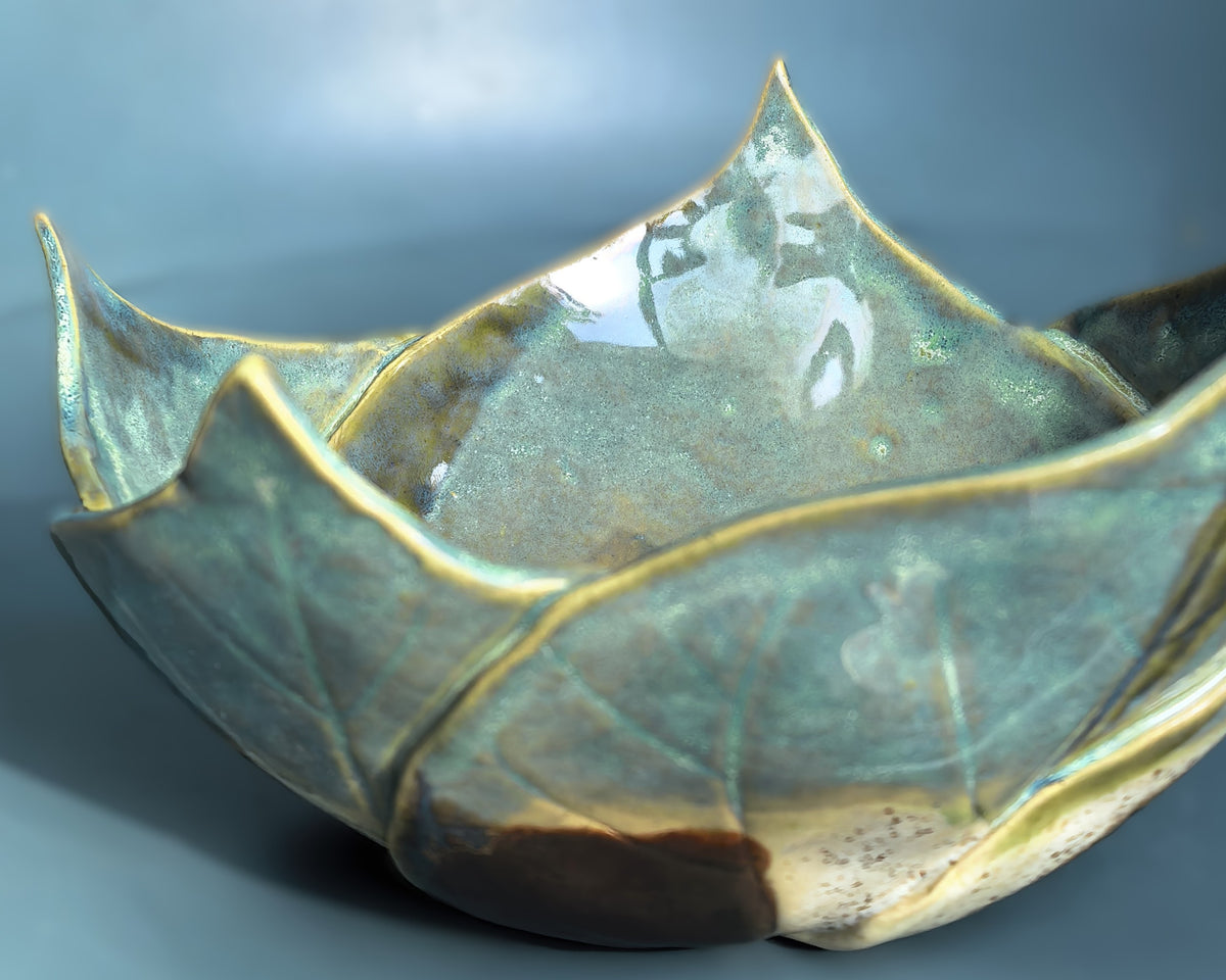 A close-up of a ceramic bowl with a lotus-petal-inspired design, featuring a glossy glaze in shades of teal, olive green, and gold, with a dark brown base.