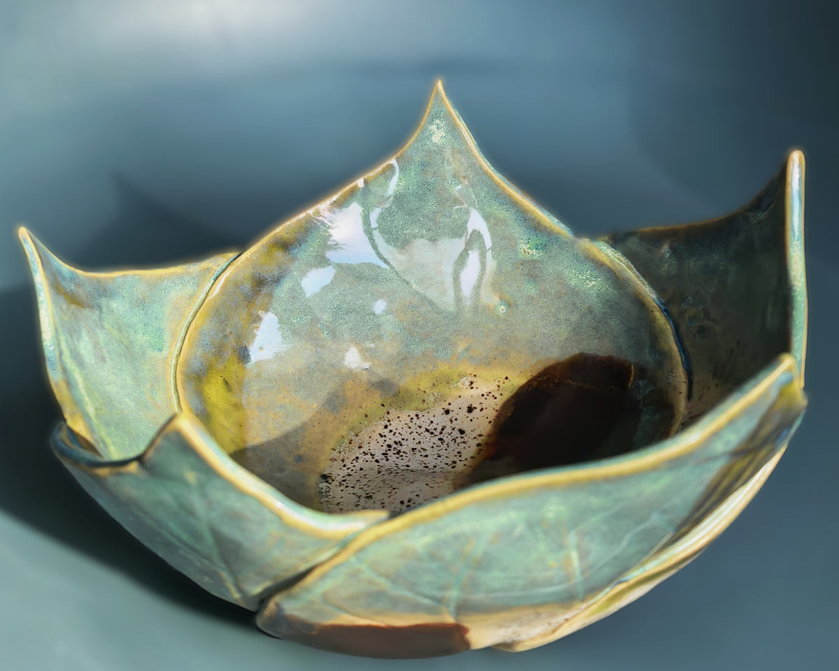 A handmade ceramic bowl with a lotus-petal shape, featuring a glossy glaze in shades of teal, green, and gold, with a dark brown speckled center and a dark brown base, set against a dark blue-gray background.