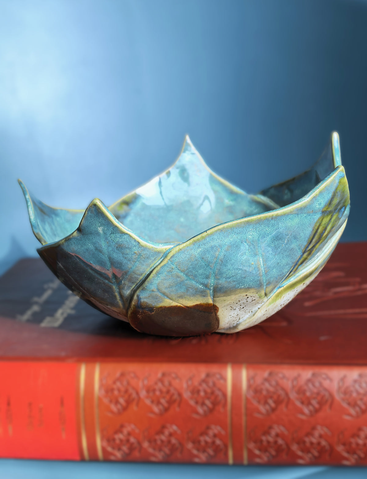 A handmade ceramic lotus-shaped bowl with a glossy teal and gold glaze, resting on top of a red vintage book against a blue background.